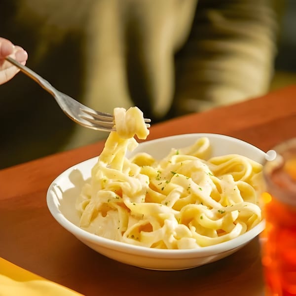 A fork lifts creamy fettuccine alfredo from a white bowl on a brown table.