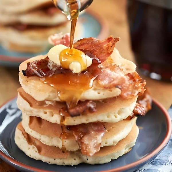 Maple syrup being poured over a stack of pancakes with bacon and butter on a blue plate.